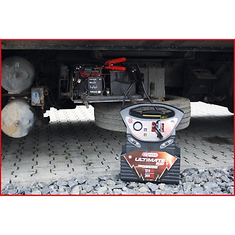 Ein Starthilfegerät steht vor einem LKW. Rote Kabelklemmen sind mit der Batterie verbunden.